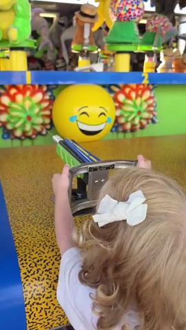 A young girl with blonde curly hair and a white bow is playing a carnival game. She is holding a toy gun that is mounted on a stand. The gun has green, blue, and yellow accents. The girl is aiming at a large yellow inflatable ball with a laughing emoji face printed on it. The ball is positioned behind a row of colorful floral decorations. In the background, there are shelves filled with stuffed animals. The game area has a yellow floor with black speckles and a blue border. The girl's left hand is on the gun's handle and her right thumb is extended upwards, indicating she might be pressing a trigger.