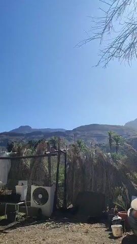 The video shows a dry, dusty landscape with mountains in the background. In the foreground, there is a structure made of wood and palm fronds, with an air conditioning unit next to it. There are also some potted plants and a car. The sky is clear and blue. The video appears to be shot during the day.