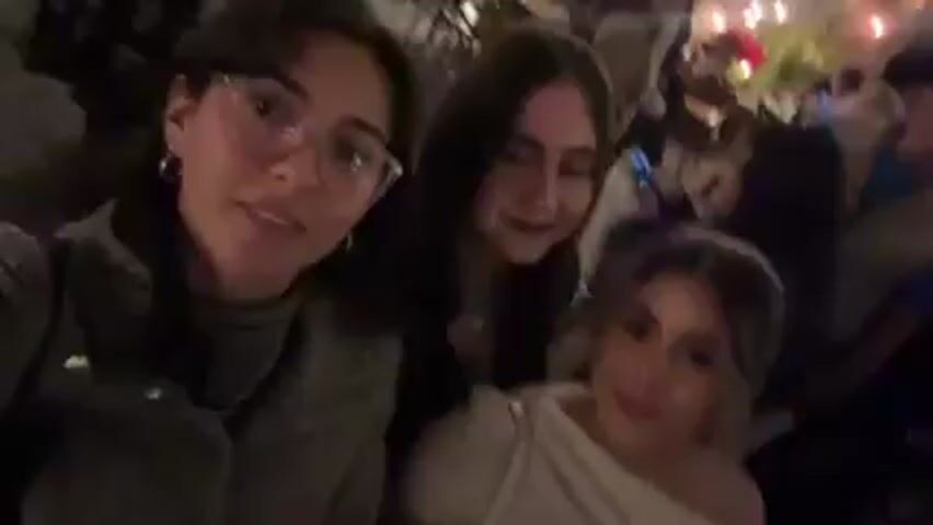 Three young women are recording a selfie video at night, standing in a crowded outdoor area. The woman holding the camera is in the foreground wearing glasses, a grey jacket, and hoop earrings. She is speaking to the camera and smiling. Behind her and slightly to her left is another woman with long dark hair. To the right of the woman holding the camera, another woman with long hair is seen smiling at the camera. People are milling around in the background, and decorative lights hang overhead. The atmosphere appears festive and lively.