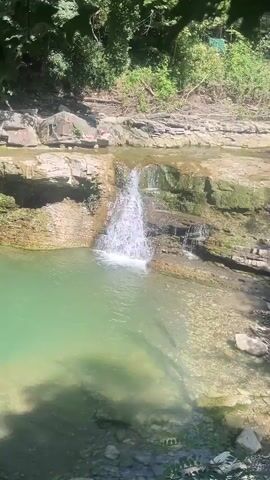 The video shows a small waterfall cascading into a clear, green pool of water. The rocks surrounding the waterfall are layered and appear to be covered in moss in some areas. Sunlight illuminates the scene, casting shadows on the rocks. In the upper portion of the frame, there are some red objects lying on the rocky ledge above the waterfall. The water is flowing from left to right, creating a white froth as it hits the pool below. The overall scene is one of natural beauty in an outdoor environment.