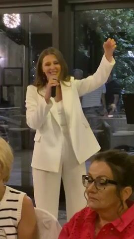 A woman wearing a white suit and a white top stands and sings into a microphone while holding her left hand up. She is in front of a group of people sitting at tables, with some foliage and lights in the background.