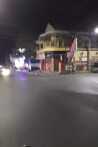 The video shows a street scene at night. The road is paved and appears wet. On the right side of the road, there is a building with yellow and blue accents, and red shutters. There are flags on poles in front of the building. Further down the street, there are businesses with illuminated signs. Several cars are driving on the road, their headlights illuminating the scene. One car in particular, a dark-colored sedan, is clearly visible as it passes by. There are also streetlights casting a purple glow on the road and sidewalk. Trees are visible along the side of the road. The overall impression is of a lively urban environment at night, with traffic and illuminated businesses.