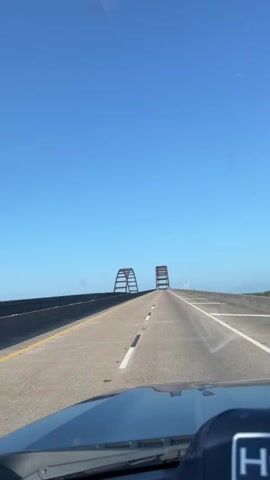 The video shows a view from the perspective of someone in a vehicle driving down a highway. The highway is an elevated road with guardrails on either side. In the distance, two large, arch-like bridge supports are visible, symmetrically placed on either side of the road ahead. The sky is a clear, bright blue with no clouds. The road surface appears to be asphalt, with white lane markings. The dashboard and windshield wipers of the vehicle are partially visible in the foreground. The overall environment suggests a sunny day, possibly over water or a low-lying area, given the elevated nature of the highway and the distant horizon.