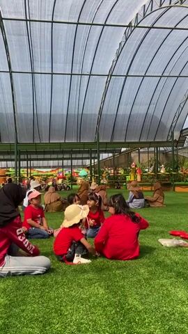 A group of children and adults sit on artificial grass under a large white arched translucent roof. The children wear red shirts. Some adults wear hijabs. The background shows more people and colorful decorations. In the first shot, a woman in a red shirt and black hijab sits on the left, facing other children. In the second shot, a woman in a striped shirt and jeans stands, facing the children, some of whom are in the same red shirts.