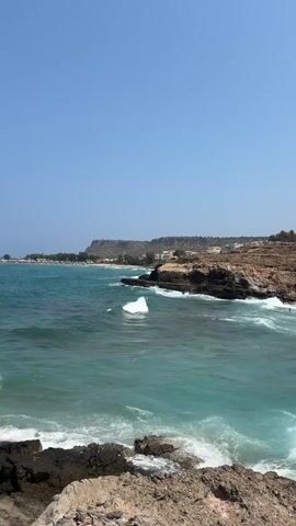 A vertical, wide shot captures a clear blue sky above a vibrant turquoise ocean. Waves crash against dark, rocky shorelines in the foreground and midground. In the distance, a beach with scattered umbrellas and buildings is visible along a gentle coastline. The overall scene depicts a sunny day at a rocky beach.