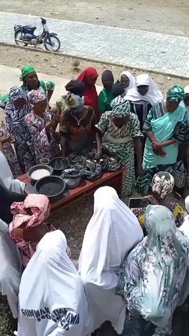 A group of about 15 women stand around a small wooden table outdoors. Several of the women are wearing bright and colorful patterned outfits and headscarves; others are wearing white or light colored full-body coverings. On the table are several bowls containing what appears to be rice, black grains, and some items wrapped in black plastic bags. The women are looking at the table and appear to be discussing or examining the contents. In the background, there's a motorcycle parked on a paved area next to a sandy or dirt space. Some writing is visible on the back of one of the white coverings. The scene appears to be taking place in a public or semi-public area, possibly in a village or town setting.