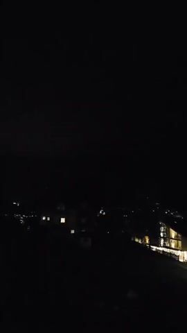 The video captures a dark night scene, likely a mountainous area with scattered buildings illuminated by artificial lights. The overall impression is one of a rural or semi-rural setting. The sky is mostly black, with some faint lights visible in the distance suggesting a settlement. There are also some illuminated structures that appear to be houses or possibly hotels, with warm light emanating from their windows. A faint, purple-blue flash of lightning briefly illuminates the silhouette of a mountain range in the middle of the frame, indicating a thunderstorm is occurring. The video is very dark, and the lightning flash is the most dynamic element. No people are visible.