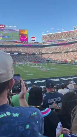 The video shows a crowd of people watching a football game at Levi's Stadium. People are holding up their phones to record the game. The scoreboard shows a live feed of the game, as well as game statistics. A large Pepsi logo is also visible. The sky is clear and sunny. Most fans are wearing casual attire. One fan wears a Seattle Seahawks Chancellor Jersey, #31.