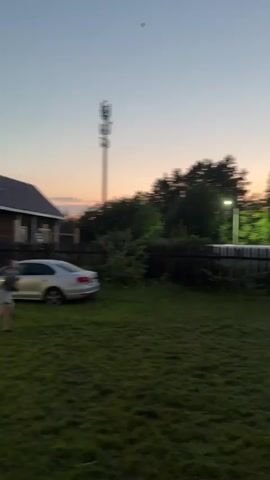 Two young females are in a grassy yard at dusk. One female is in the foreground looking up and throwing something into the air, a kite, which is visible in the sky. The other female is standing further back in the yard, also looking up. There is a fence, trees, a cell tower, a house, and a car in the background.