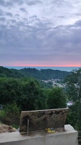 A video captures a wide panoramic view of a city nestled among lush green hills, with the sea stretching out to the horizon under a dramatic sunset. The sky is a canvas of vibrant orange and pink hues, contrasting with the soft blue and grey clouds. The city's buildings are scattered throughout the landscape, with some lights twinkling on, indicating the approaching evening. The foreground features a dirt patch with some construction debris, including a yellow object and some wooden planks. The camera slowly pans to the right, revealing more of the rolling hills and the distant city lights. The overall ambiance is peaceful and picturesque, highlighting the beauty of nature and the urban landscape blending together.