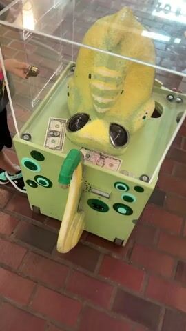 A yellow, alien-like creature sits inside a clear plastic box with a light green base. Several one-dollar bills and some coins are scattered on the base, in front of the creature. The creature has large, dark eyes with a crescent moon reflection. A green lever is attached to the side of the box. A child's arm is visible in the upper left corner, holding a golden knob. The box is placed on a brick-like surface.