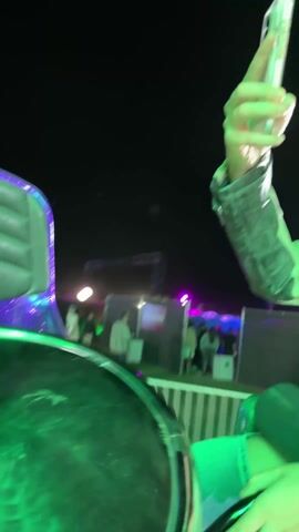 A person is riding a fairground ride. The person is holding a smartphone, apparently recording a video of the ride. Several other fairground rides can be seen in the background, turning rapidly. Other fairground visitors are visible. The scene is at night and is well-lit with colorful spotlights.
