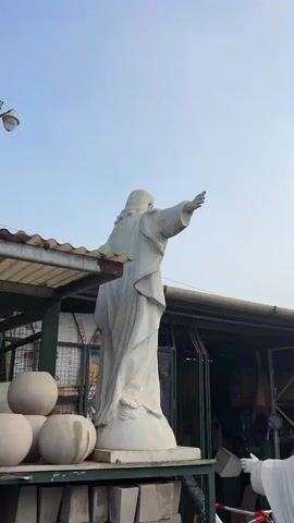The video shows a white statue of Jesus Christ with open arms, standing on a platform. The statue is positioned outdoors, possibly at a garden center or similar establishment. Behind the statue are stacked round, pale planters and a corrugated metal structure with a light fixture attached. The sky is visible in the background. The camera moves, slowly panning to reveal more of the statue's face and upper body.