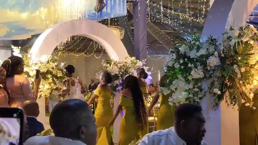 The video shows a group of people at a wedding reception.  In the foreground, people are dancing, and a child in a suit stands near a woman in a pink dress. Behind them, the bride, in a white dress, is dancing with several women in yellow dresses.  There are white floral arrangements on either side of the dance floor.  A large, white, arch-shaped structure stands behind the dance floor, and string lights hang from the ceiling. People are watching and taking pictures.  A man is seen taking a video of the scene.