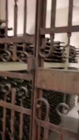 The video shows a wrought-iron gate leading to a wine cellar. The gate is rusted and has decorative curls and bars. Behind the gate, shelves are stacked with wine bottles. The bottles are various shades of green and are arranged horizontally. The room is dimly lit and appears to be made of stone. There is a faint sound of voices in the background, suggesting the presence of people.