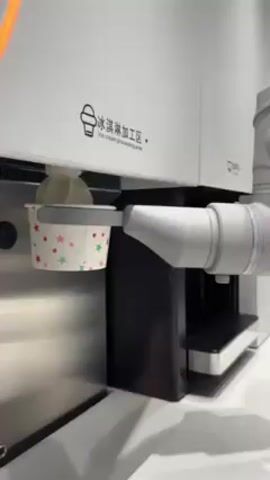The video shows a machine dispensing ice cream into a small paper cup. The machine is white with a black and stainless steel accent. There are the Chinese characters “冰淇淋加工区” and below that the English, “ice cream processing area” on the front. A gray robotic arm holds the paper cup up to a dispenser. A swirl of white ice cream is dispensed into the cup. The paper cup has a pattern of red, green, and pink stars on it, as well as a gray scalloped edge on the top.