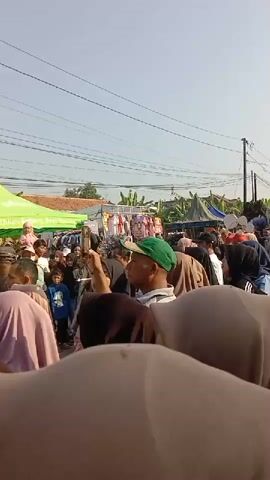A video displays a street market scene with several stalls and a crowd of people. A green tent with white lettering is visible on the left side of the frame. Behind it, clothing is displayed on racks. Several individuals, including children and adults, are milling about. Some people are wearing headscarves. In the foreground, several people are partially visible, obscuring the view of the market. The sky is clear and bright, suggesting it is daytime. The environment appears to be a residential area with houses and trees in the background. The overall atmosphere seems to be of a lively gathering or event.