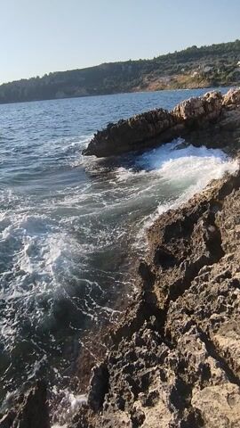 This media file shows a close-up view of waves crashing against rocky cliffs on the coast of Karaburun, Turkey. The sun is shining brightly, indicating it is daytime. The water is a deep blue, and the waves are creating a foamy white spray as they hit the rugged, dark rocks. The rocks have a porous texture and appear to be ancient. In the background, a lush green hillside slopes down towards the sea, suggesting a natural, unspoiled environment. The overall mood is one of natural beauty and the raw power of the ocean. There is no visible text in the media file.