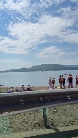The video shows a view from a moving vehicle, looking out over a body of water. On the shore, there is a black metal guardrail, followed by a dirt path. Beyond the path, there is a large body of water with a distant, tree-covered hill. There are a few small boats on the water, and a few people are visible on the shore, some sitting and some in the water. A sign is posted on the shore. The sky is blue with scattered white clouds. The vehicle passes a black SUV, then a white sedan behind a black metal fence, then a brick building with a round tower.
