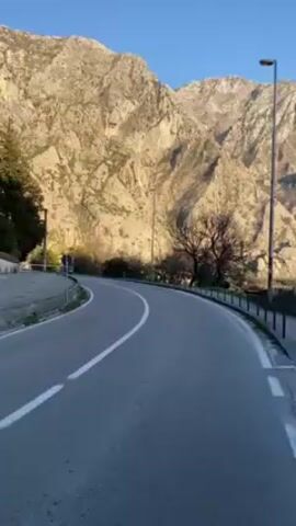 The video shows a winding road with a mountain backdrop. The road is paved and has white lane markings, as well as a sidewalk with a metal fence on one side. Several bare trees dot the scene. The mountain is large and rocky, and lit by the sun. The sky is a clear blue. A tall street light stands near the road. In the distance, a body of water can be seen. The camera is moving along the road, giving the viewer a sense of travel.