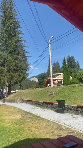 The video shows a daytime outdoor scene in a mountainous, forested area. A tall evergreen tree dominates the left side of the frame. Power lines and a utility pole are visible in the center. In the background, there is a building with a red roof and an elevated walkway, and more trees and a rocky mountain face. A gravel path runs through a grassy area. Several people are scattered throughout the scene, some walking, some sitting. One person is standing near a small red gazebo with a picnic table. The view is framed by a wooden structure in the foreground, suggesting the video is being filmed from under a covered area.
