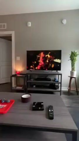 The video shows the interior of a living room, transitioning to an exterior view. Initially, the camera focuses on a living room setup featuring a TV displaying a vibrant scene, a grey TV stand, and a coffee table with various items. The room is decorated with plants, a skateboard mounted on the wall, and modern furniture. The camera then pans towards a window offering a view of a courtyard. The courtyard contains a swimming pool surrounded by buildings with blue accents. People are visible around the pool, lounging and engaging in leisure activities. The scene is bright and airy. Music is playing in the background.