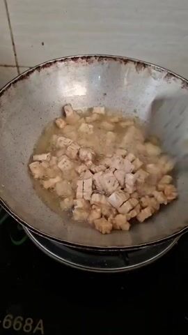A metal wok filled with oil sits on a stovetop. Small, light brown, cubed pieces of food are being stirred in the hot oil with a metal slotted spoon. The food is in various stages of frying. The wok is on a black electric stovetop with green accents. The background appears to be a tiled wall.