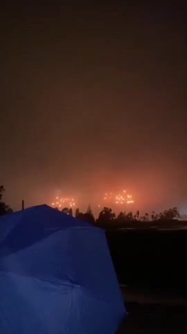 The video captures a nighttime scene with an open blue umbrella in the foreground, partially obscuring the view of a distant fireworks display. The fireworks are erupting in a hazy, reddish-orange glow against a dark, foggy sky. The silhouettes of trees and possibly some structures are visible between the umbrella and the fireworks. The overall atmosphere is dark and suggests it might be raining due to the presence of the umbrella.