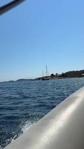 The video begins with a view of choppy blue water from the perspective of a boat, with the bow of the boat visible in the foreground. A sailboat with two masts is anchored offshore near a rocky coastline. Buildings, including what appears to be a restaurant with tables and chairs and a windmill with a red roof, are situated on the hillside behind the sailboat. The sky is clear and blue. The video pans slightly to the right, revealing more of the coastline and the buildings.