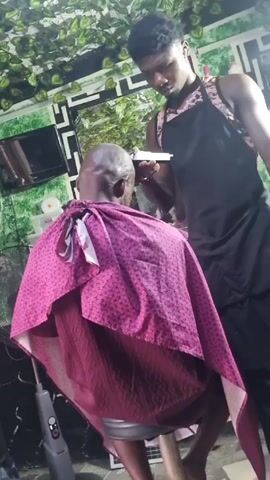 Here is a concise description of the scene:

Inside a barber shop, a person sits on a metal stool with a maroon and pink cape, facing away from the camera. The person's head is bald, showing some stubble. A barber, wearing a printed tank top and black apron, stands behind the seated person, using a pair of clippers on the person’s head. A mirror on the wall reflects the scene. The shop is decorated with artificial ivy, and there's a television, a fan, and various barber supplies visible in the background.