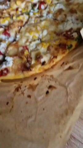 A top-down view reveals a pizza sitting on brown parchment paper inside a brown paper bag. The pizza is cut into slices. The toppings consist of corn kernels, red peppers, and what appears to be white cheese. A hand reaches into the shot and grabs a slice of pizza. A white plate is visible in the upper left corner.