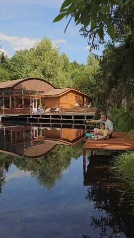 A man and a child sit on a wooden dock extending into a calm body of water. They appear to be fishing, with a fishing rod visible. The water reflects the surrounding trees and a wooden building with a thatched roof in the background. The building has large windows and appears to be a lodge or resort. Lush green foliage frames the scene. The sky is partly cloudy.