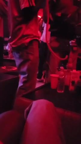 The video is shot from a low perspective, showing a dimly lit, possibly indoor, social gathering. The lighting shifts in color from red to blue to green. Several people are visible standing and moving around, but they are mostly out of focus. A table in the foreground has cups, water bottles, and other containers on it. A portion of what appears to be a person's leg, covered with light-colored fabric, is also visible in the foreground. The overall atmosphere seems relaxed and social.