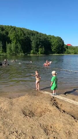 The video shows a summertime scene at a lake. Several people are swimming in the water, and a group is on an inflatable raft. Two young boys stand on a sandy shore near a log extending into the water. The boy closest to the camera wears striped swim trunks and is facing the water, while the other boy, dressed in a green t-shirt and a light-colored hat, points toward the lake. The environment includes a backdrop of lush green trees and a clear blue sky. A red roof is visible through the trees on the far right.