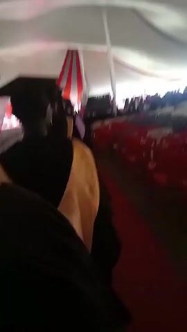 The video shows the inside of a large tent. There are rows of chairs, some red and some white with red sashes. A person in a graduation gown walks down a red carpet in the aisle. A large screen and decorative flags are visible at the front of the tent. The tent has a white ceiling and red accents.