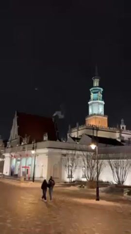The video shows a couple walking along a snow-covered, paved path in front of two buildings at night. The building on the left is white with a red roof and is decorated with Polish flags. Smoke is visible rising from a chimney on its roof. The building on the right is a tall, ornate structure with a spire. It is illuminated with white and yellow lights. A street lamp stands on the path in front of the couple. The sky is dark.