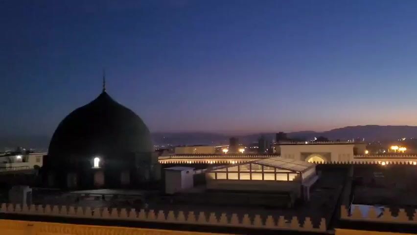 The video shows a nighttime cityscape with a large, dark dome in the foreground, possibly part of a mosque or similar structure. The dome has a single illuminated window. Behind the dome is a rooftop with several structures, including a small building with a glass roof illuminated from within. Decorative lights line the edges of the rooftop. In the distance, mountains are visible on the horizon, with city lights scattered across the landscape. The sky is a gradient of dark blue to orange, indicating either dusk or dawn. The overall scene is still and quiet.