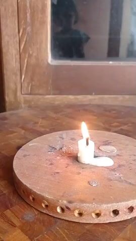 A white candle is lit and placed on a round, wooden surface with holes around the edge. The candle is melting and wax is pooling around its base. In the background, a wooden cabinet with a glass pane is visible. A blurred reflection of a person is seen in the glass. The video is approximately 5 seconds long and appears to be a short clip of a birthday candle being lit or about to be blown out.