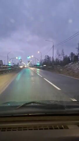 The video shows a view from inside a vehicle driving down a wet road, likely at dusk or dawn. There are overhead traffic lights displaying green signals and pedestrian crossing signs above them. Streetlights illuminate the road. Some buildings and trees are visible to the side of the road. There are few other cars visible. The sky is overcast.