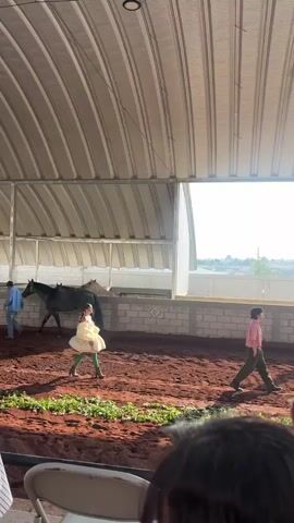 The video shows an indoor dirt riding arena with a corrugated metal roof and block walls. Outside the arena is a view of a rural landscape under a bright, sunny sky. The foreground view is over the heads of spectators seated on chairs. A girl wearing a yellow dress and green leggings is performing alone in the dirt arena. She is walking in a circle and turning. A man wearing pink and jeans is also walking, near the edge of the arena. Further along the edge, a man in a baseball cap is leading a horse. A second horse is visible behind the first one. Another person wearing a light blue jacket and white pants walks into the arena.