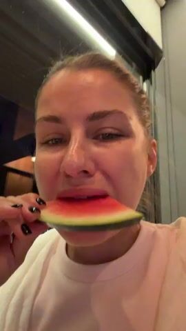 This is a close-up video of a young woman eating a slice of watermelon. The woman is of Caucasian descent and has her hair pulled back. She is wearing a light-colored t-shirt. Her fingernails are painted black. The watermelon slice is large and appears to be fresh, with a bright red interior and a green rind. The woman is holding the watermelon slice with her right hand and is in the process of taking a bite. Her mouth is slightly open, and her cheeks are puffed out as she chews.

The background of the video is out of focus, but it appears to be an indoor setting with some lighting fixtures visible overhead. There are also some dark shapes in the background that could be furniture or architectural elements. The lighting suggests it might be evening or a dimly lit indoor space.

The woman's facial expression is difficult to discern due to the close-up nature of the shot and the focus being on her eating. However, the act of eating watermelon is generally associated with enjoyment and refreshment, especially in warm weather.

There is no discernible text visible in the video. The context provided indicates the location is Žunovnica, Bosnia and Herzegovina, which could imply a casual dining or social setting. The overall impression is a candid moment of someone enjoying a refreshing snack.