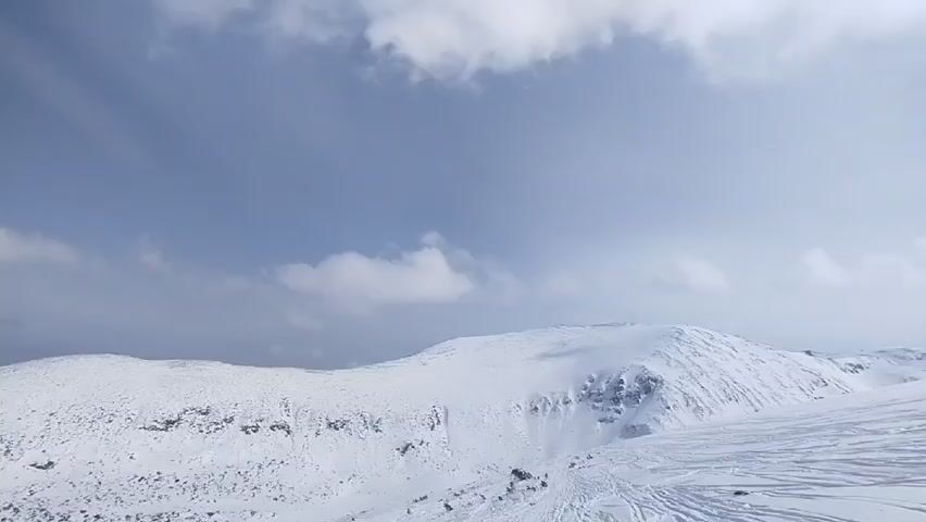 The video shows a snowy mountainous landscape under a cloudy sky. The mountains are covered in snow, and there are visible tracks on the snow. A valley is visible between the mountains. The sky is blue with scattered white clouds.
