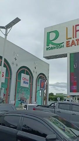 The video shows a sign for Robinson Lifestyle Eat Shop Play. The sign is large and colorful, with the word "Robinson" in green on the left, and "Lifestyle Eat Shop Play" in various colors on the right. Below the main sign is a large digital billboard displaying Thai text and graphics. To the left of the sign is a building with arched windows and a sign that reads "Office Mate." In the foreground, several cars are parked in what appears to be a parking lot. The sky is overcast, and trees and a gas station sign are visible in the background.