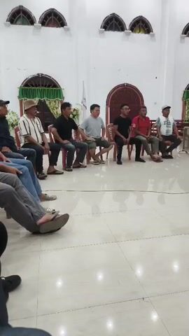 The video shows a group of men sitting in chairs in what appears to be a church hall. They are arranged in a line facing a keyboard player who is seated and playing an electronic keyboard. The room has high ceilings with arched windows and decorative trim. There are curtains on the windows and some floral arrangements. The floor is tiled and reflective. The men are casually dressed. The keyboard player is wearing a t-shirt and pants and is focused on the keyboard. The overall atmosphere seems to be of a rehearsal or a small performance.
