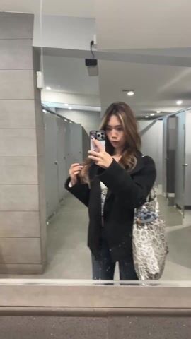 A young Asian woman is taking a selfie in a public restroom, reflected in a large mirror. She is wearing jeans, a white shirt, and a black coat, and has a large silver and white leopard print tote bag slung over her shoulder. Her hair is highlighted. The restroom has gray tile walls and stalls visible in the background. The video focuses on the woman as she adjusts her phone and poses.