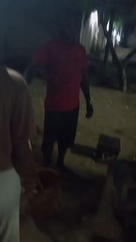 The video shows an outdoor gathering at night. Two men are visible, one wearing a red shirt and dark pants and the other wearing a pink shirt and dark pants. A partially visible third person is seen wearing a white garment. The two men in the foreground appear to be serving food from a pink bucket, possibly at a small table or stool. The area seems to be a yard or outdoor space with trees and a building in the background. Illumination is provided by what looks like outdoor lighting. The video is shaky, and the subjects are not sharply in focus.
