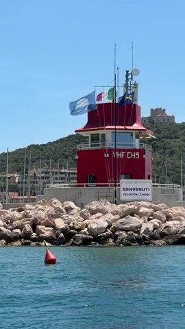 A bright red lighthouse with a white base stands on a rocky jetty. The lighthouse has a red roof with white trim and several antennas on top. Flags are flying from the top of the lighthouse, including what appears to be the Italian flag. A sign on the lighthouse reads "BENVENUTI VELOCITA' 3 NODI". In the foreground, blue water with ripples is visible, and a red buoy is floating in the water to the left. In the background, a green, tree-covered hill rises behind a town with buildings and more flags. The sky is clear and blue.