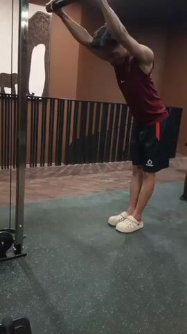 A young man wearing a maroon tank top and black shorts with a red stripe is performing a cable pushdown exercise in a gym. He is standing upright, holding a straight bar attachment connected to a cable machine with an overhand grip, his arms extended above his head. He bends his elbows, lowering the bar behind his head while keeping his upper arms stationary. He is wearing light-colored clogs. The gym has a dark gray rubber floor and brown walls. In the background, another person wearing a dark shirt and brown pants is partially visible using exercise equipment.