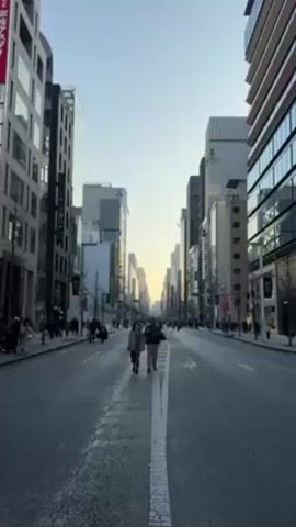 The scene shows a long shot of a wide street flanked by tall buildings in an urban environment, likely a city.  A couple is walking hand-in-hand down the middle of the street towards the viewer.  Several other pedestrians are walking along the sidewalks on either side of the street. The sky is a clear, pale blue.  A white line runs down the center of the street. The buildings vary in design and height. Some of the buildings have visible signage.  The overall mood is calm and peaceful.