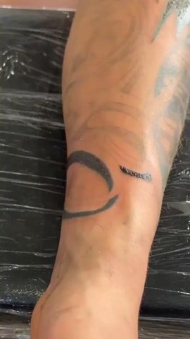 The video shows a close-up view of a person's leg with a tattoo being worked on. The leg is partially covered in plastic wrap, and the tattoo appears to be a large, dark design. A tattoo stencil, featuring a skeleton holding a pocket watch, is also visible on a piece of paper. The lighting suggests the video was taken indoors, possibly in a tattoo studio.